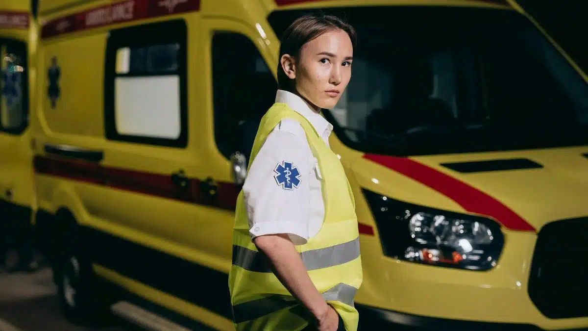 A female paramedic in a reflective vest stands beside a yellow ambulance at night.