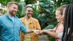 A diverse team of adults enjoying a fist bump gesture in an indoor greenhouse setting.