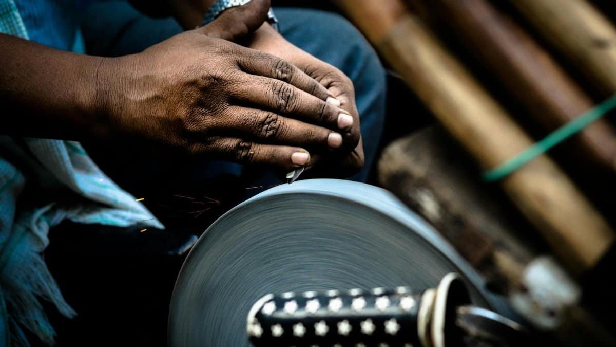 Hands sharpening a knife against a rotating grindstone in a market setting.