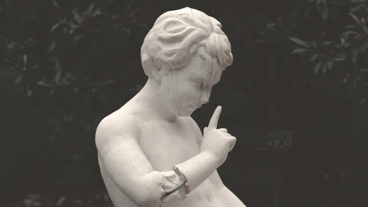 A classical marble statue of a child in a gesture of silence, surrounded by lush foliage.