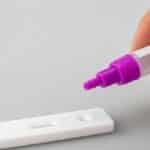 Close-up of a hand applying liquid on a rapid test kit, against a white surface.