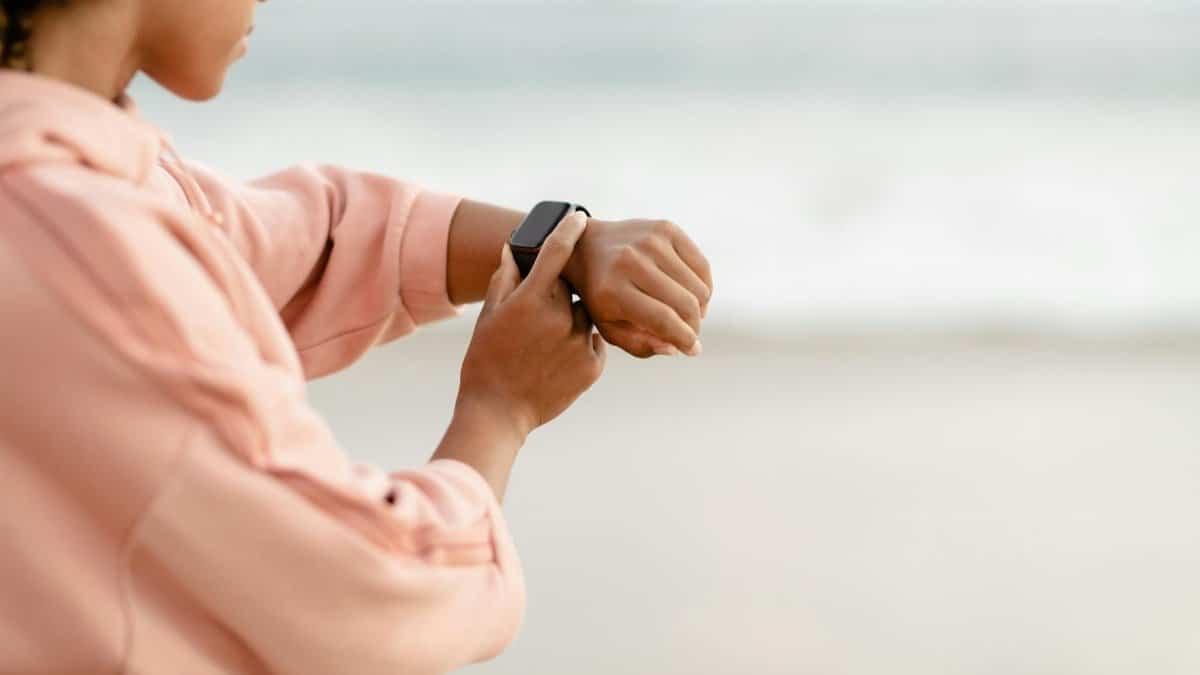 Woman wearing pink hoodie checks smartwatch by the ocean, focused on time tracking.