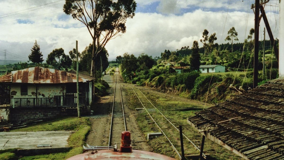 Train tracks through a rural landscape in Ecuador with cloudy sky and old houses.