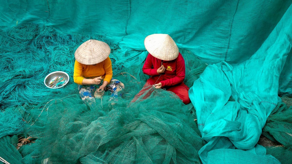 Two women in conical hats mend fishing nets, showcasing traditional Vietnamese craftsmanship.