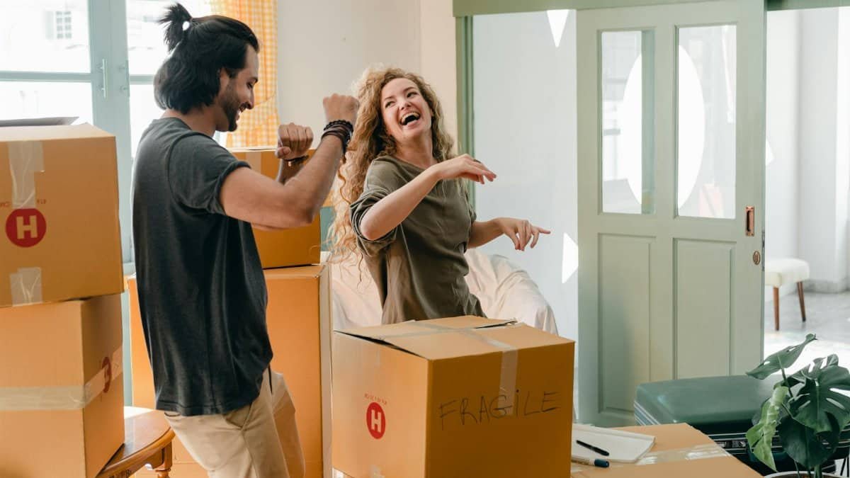 Cheerful laughing couple in casual clothes having fun and dancing together while unpacking carton boxes after moving into new contemporary apartment
