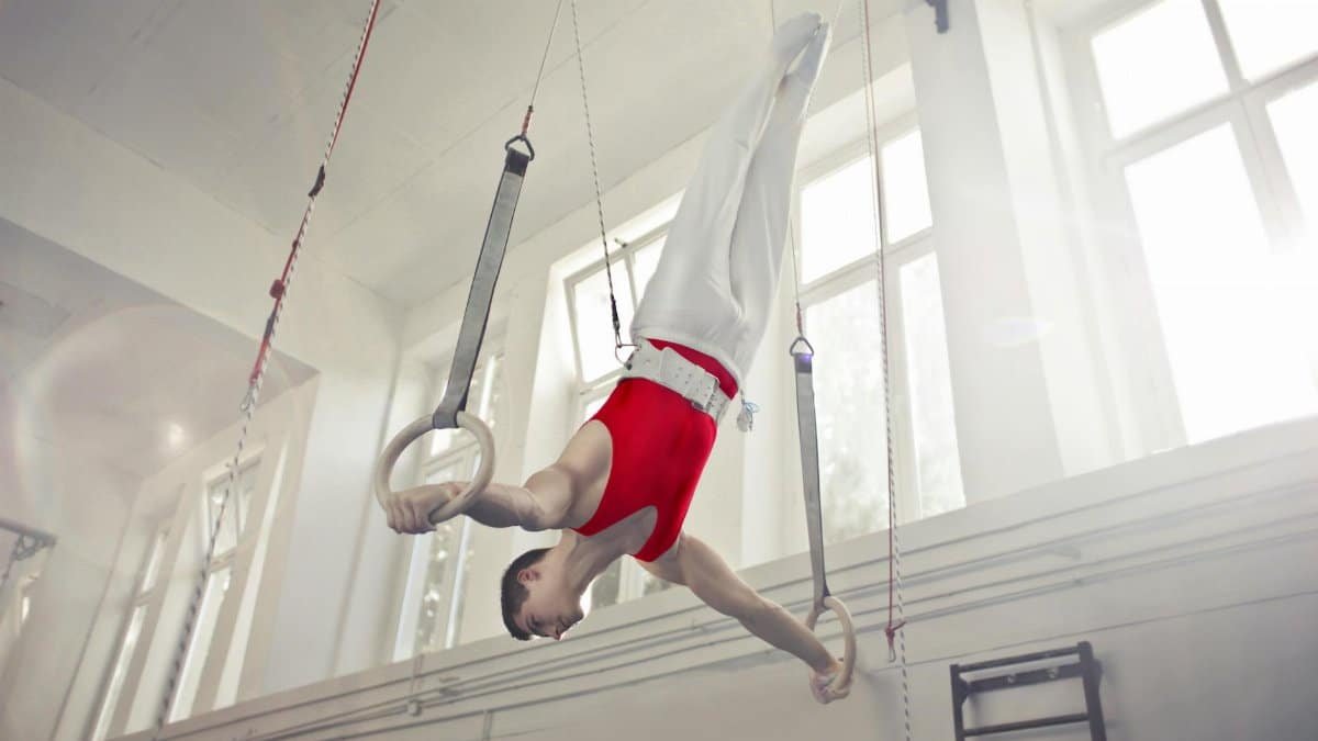 A male gymnast exercises on rings in an indoor gym, showcasing strength and flexibility.