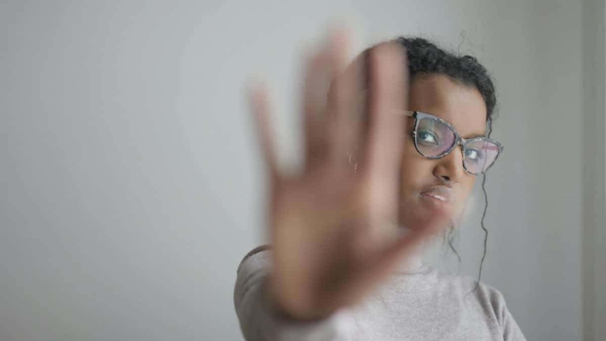 Young woman with glasses making a stop gesture against a blurred gray background.