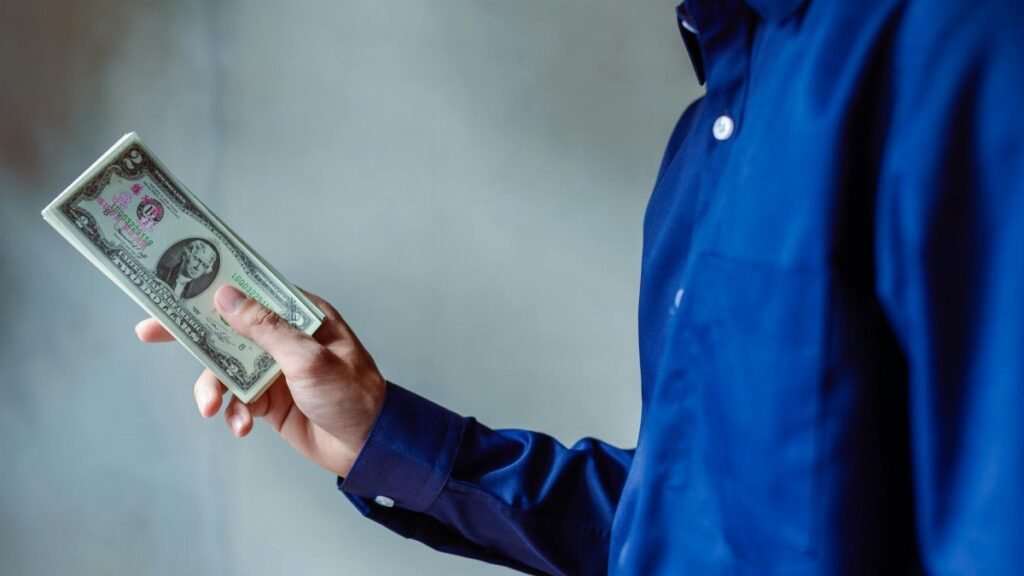 A person in a blue shirt holding a stack of US dollar bills, symbolizing wealth and finance.