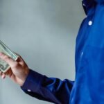 A person in a blue shirt holding a stack of US dollar bills, symbolizing wealth and finance.