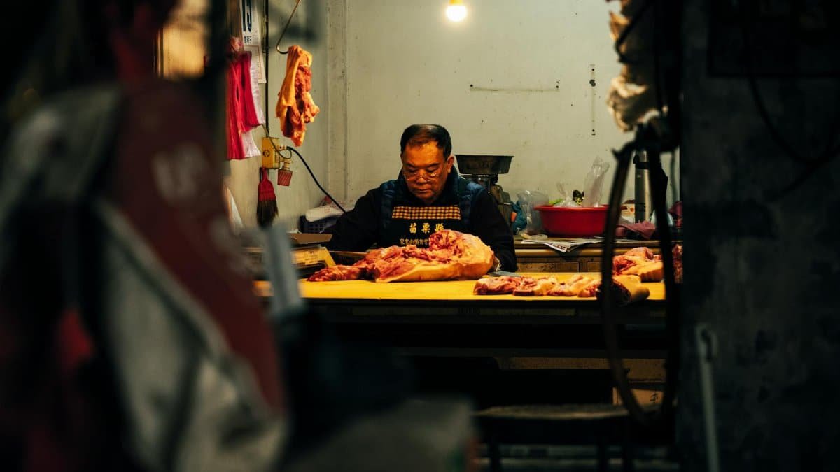 A dedicated butcher cuts meat in a dimly lit indoor setting, focused on his work.