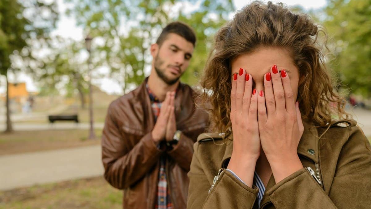 A young man apologizes while a woman covers her face, set in an outdoor park.