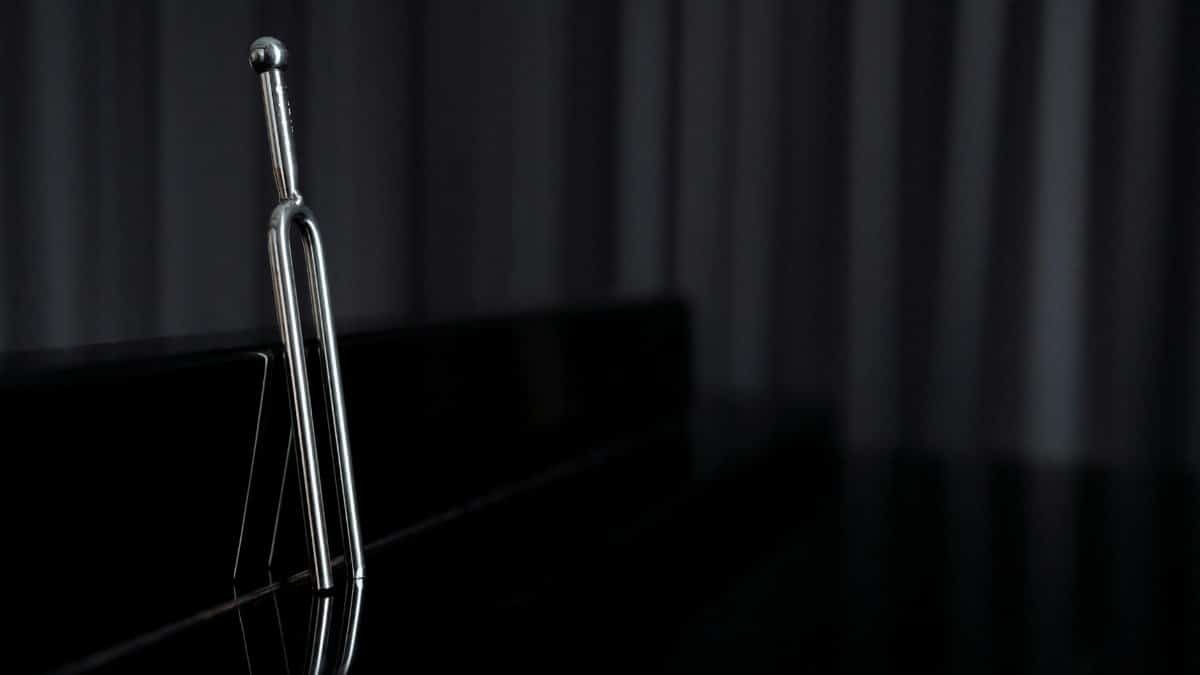 A close-up of a metal tuning fork standing on a reflective piano surface with dark background.
