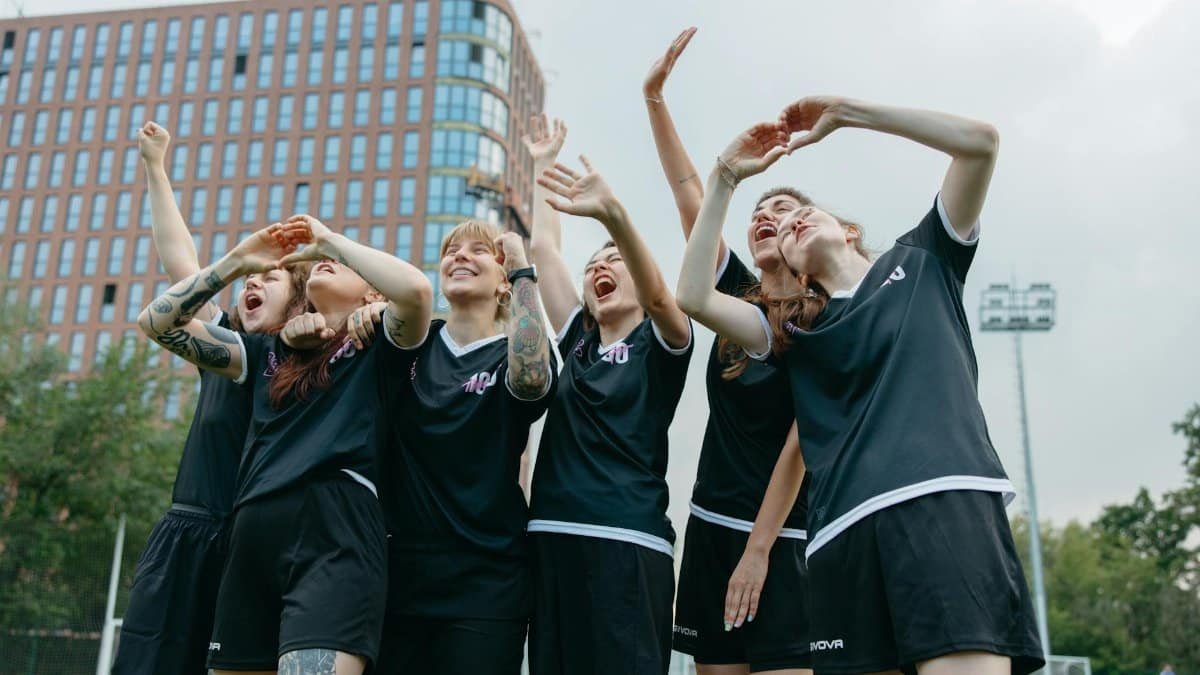 A joyful moment with women soccer players celebrating victory on an outdoor field with a building backdrop.