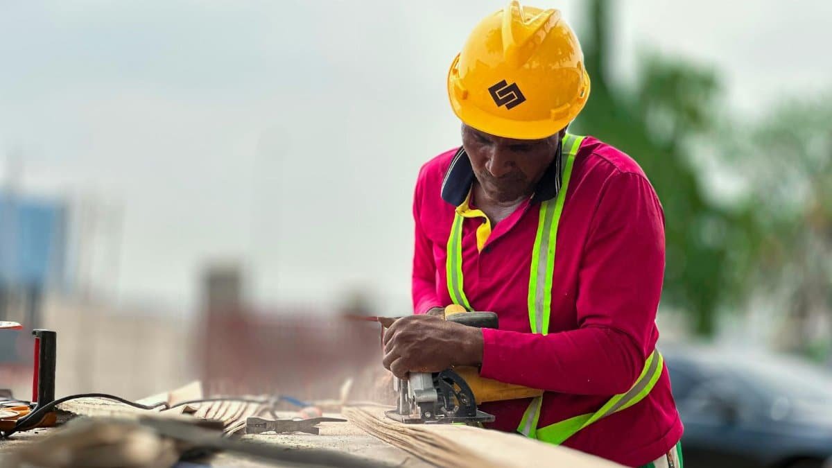 Construction worker cutting wood with a power tool outdoors, wearing safety gear.