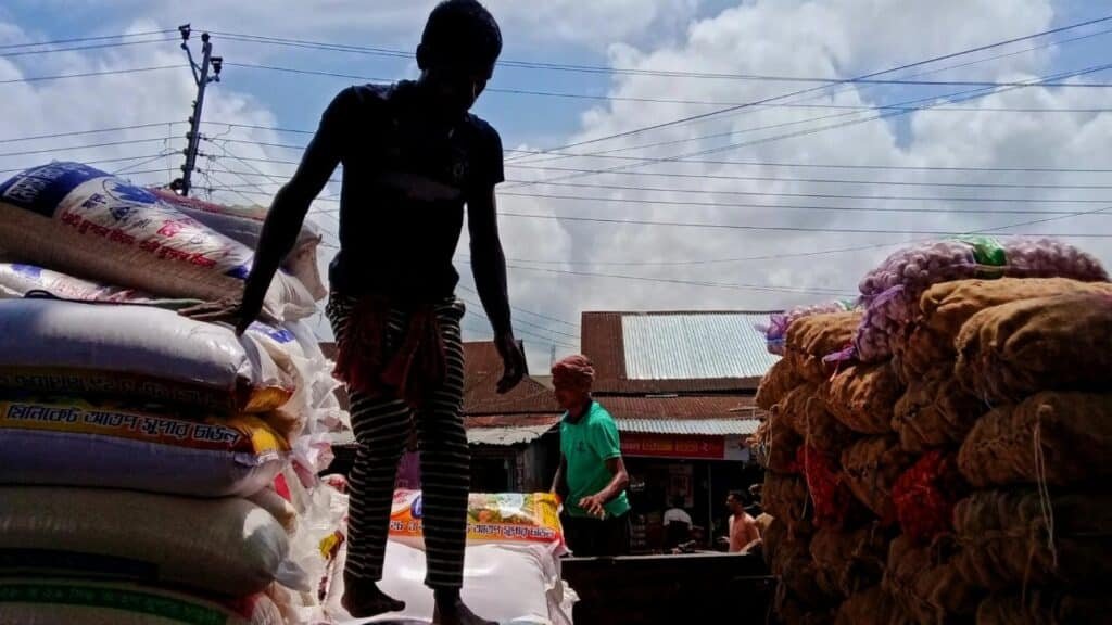 Silhouette of workers stacking large sacks at an outdoor market under a bright sky.