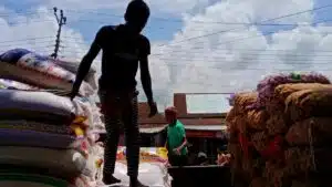 Silhouette of workers stacking large sacks at an outdoor market under a bright sky.