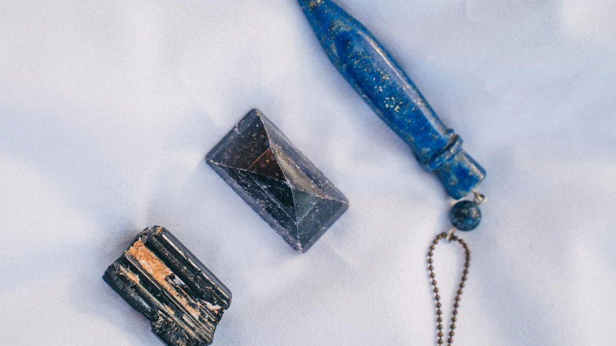 Close-up of three gemstones — black tourmaline, amethyst pyramid, and blue lapis lazuli — on white fabric.