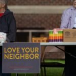 Two elderly women volunteer at a community event with a 'Love Your Neighbor' sign.