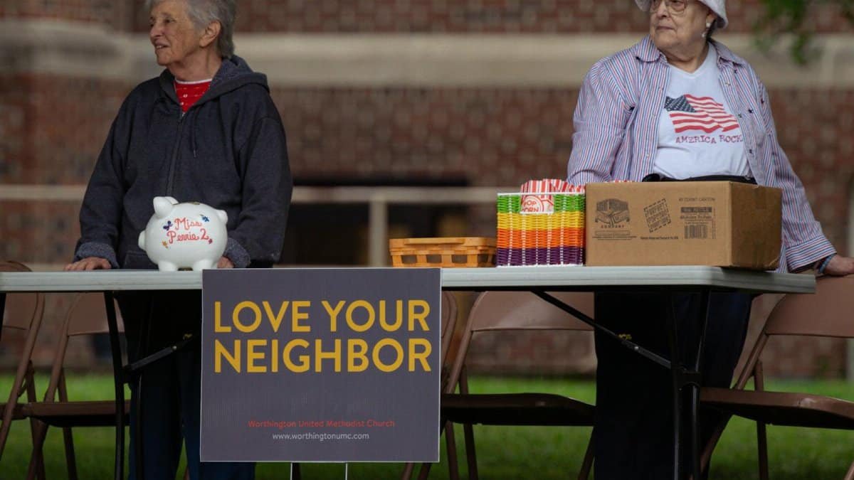 Two elderly women volunteer at a community event with a 'Love Your Neighbor' sign.