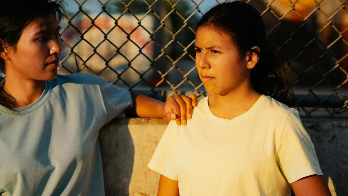 Two teenage girls engaged in a thoughtful conversation outdoors by a chain-link fence.
