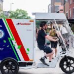 Side view of an electric delivery tricycle operated by a courier in an urban setting, promoting sustainable transit.