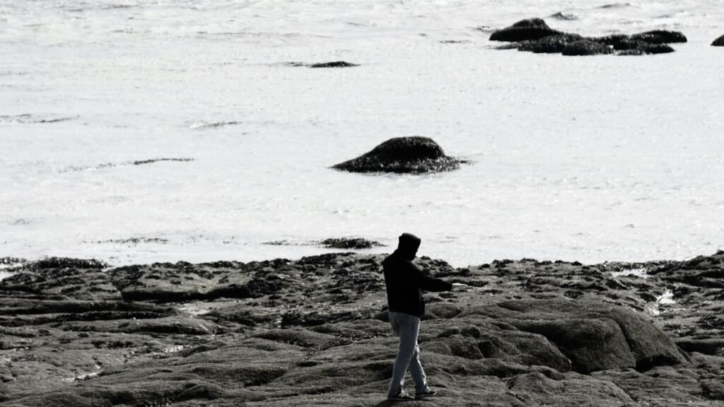 A silhouette of a man walking on a rocky beach during low tide in a black and white photograph.