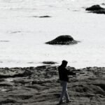 A silhouette of a man walking on a rocky beach during low tide in a black and white photograph.