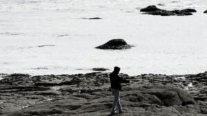 A silhouette of a man walking on a rocky beach during low tide in a black and white photograph.