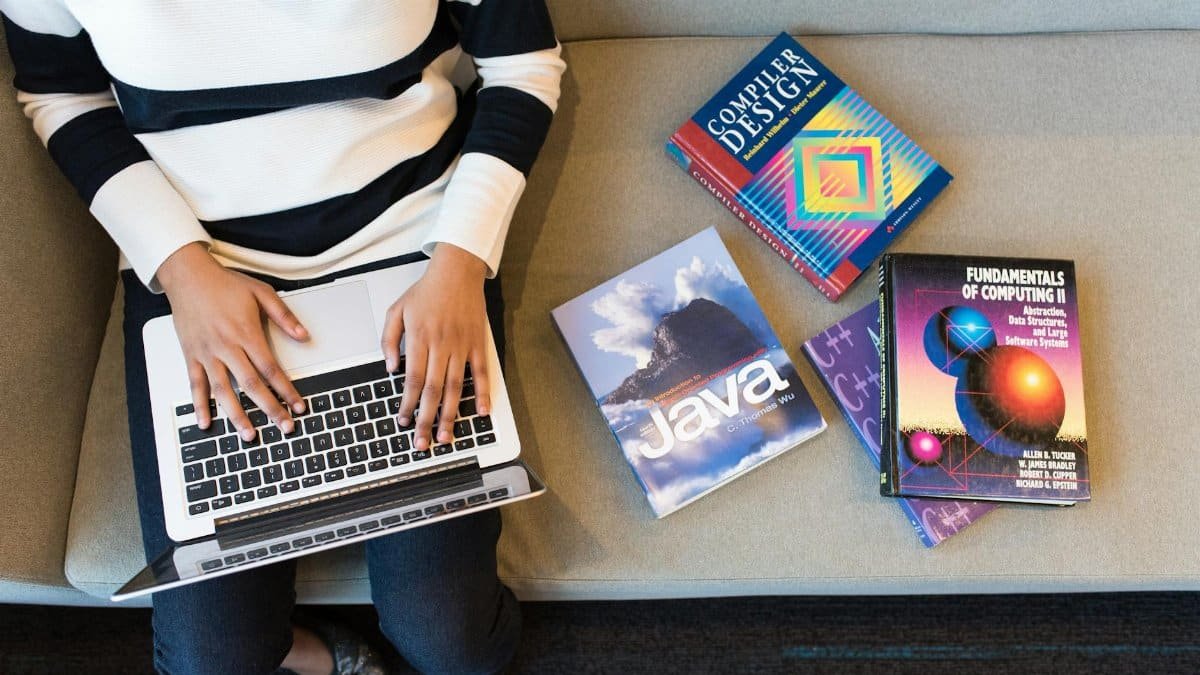 Woman using laptop on sofa, surrounded by programming books, learning coding.