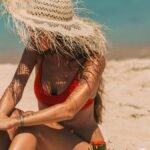 Woman in straw hat applying sunscreen while sitting on a sunny beach in summer.