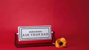A whimsical Mother's Day sign with 'Mom's Mood: Ask Your Dad' text, set against a red backdrop with a flower.
