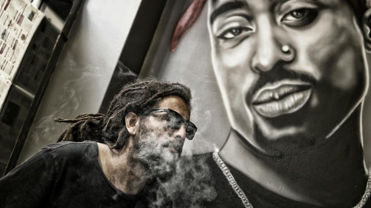 A man smoking in front of a mural featuring a hip hop artist, blending urban style and art.
