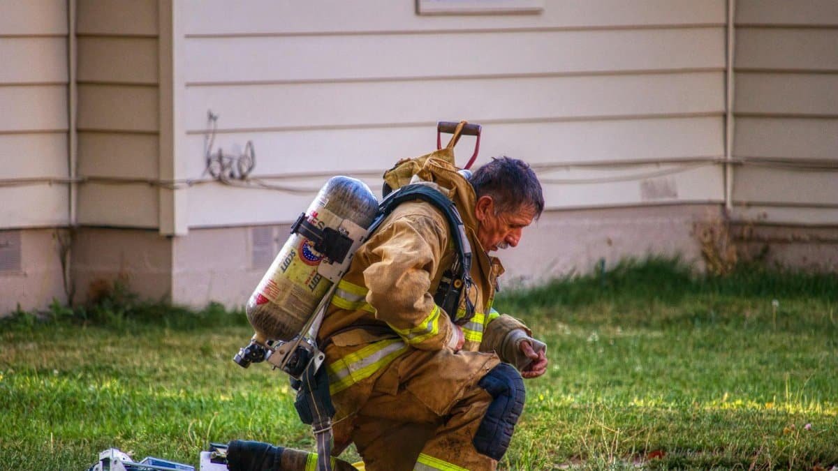 A firefighter kneels on the lawn in full gear, checking equipment beside a house.