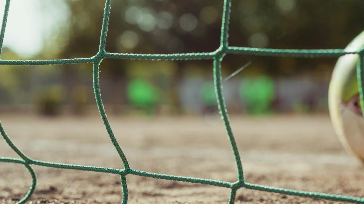 Close-up view of a soccer ball near the net on a dry outdoor field with blurred bokeh background.