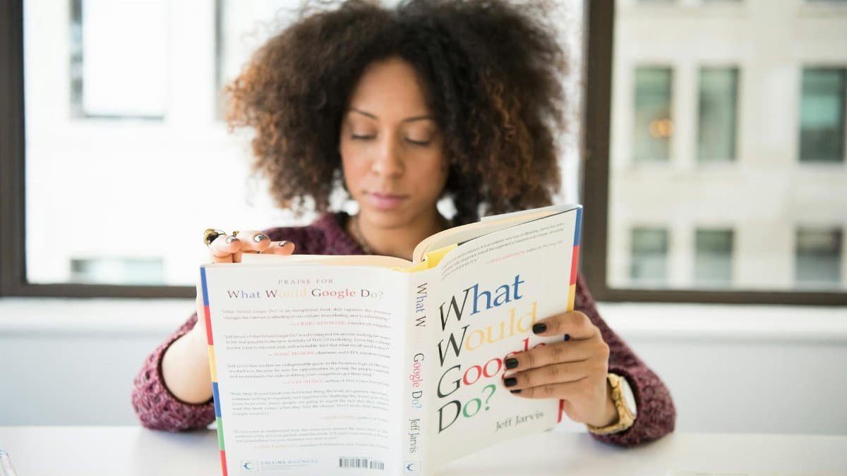 A woman reading 'What Would Google Do?' at a desk by a window in a modern office.