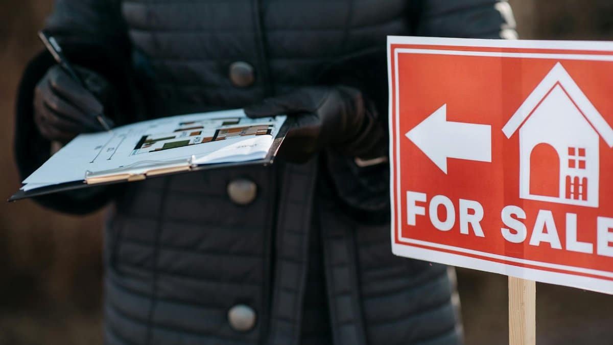 A real estate agent reviews plans next to a 'For Sale' sign outdoors, indicating property availability.