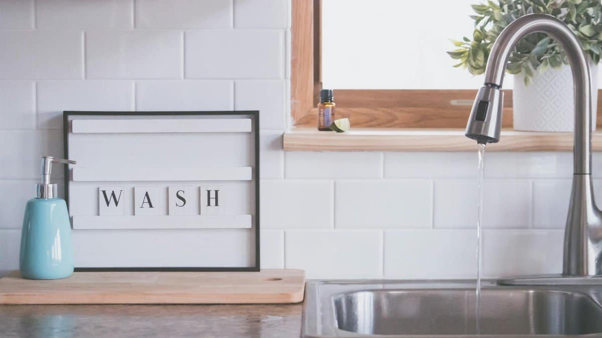 Board with word WASH placed near soap dispenser on counter in bathroom with water streaming from open faucet