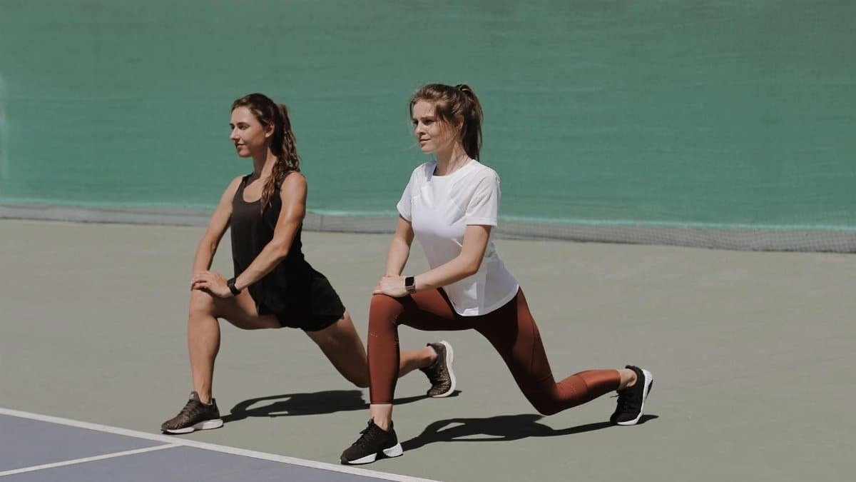 Two women perform lunges on an outdoor court, showcasing fitness and healthy lifestyle.