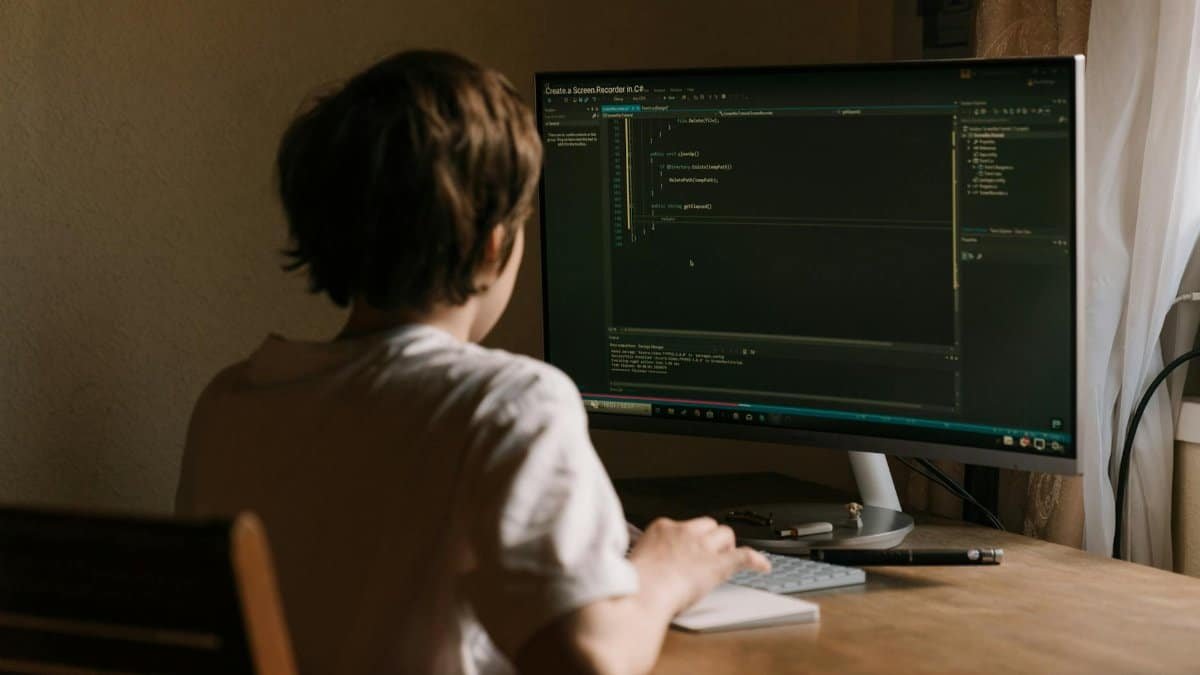 A teenager focused on coding software on a desktop monitor in a home office setting.