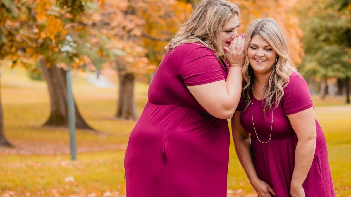 Two happy women in pink dresses gossip in a vibrant autumn park setting.