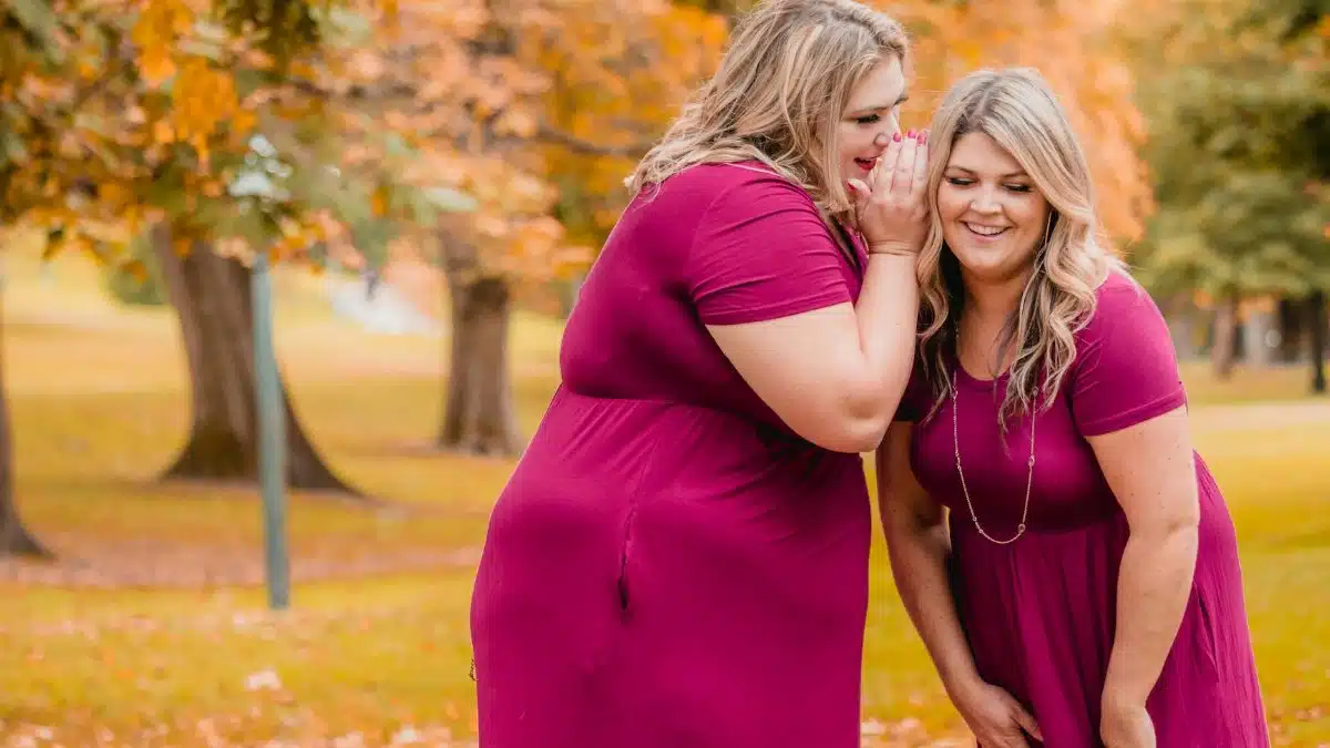 Two happy women in pink dresses gossip in a vibrant autumn park setting.