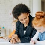 A teacher helps children with writing in a classroom setting, showcasing a diverse learning environment.