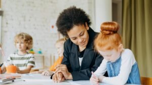 A teacher helps children with writing in a classroom setting, showcasing a diverse learning environment.