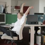 Side view of cheerful female employee in formal outfit sitting on office chair with crossed legs on desk and stretching while resting during work with closed eyes