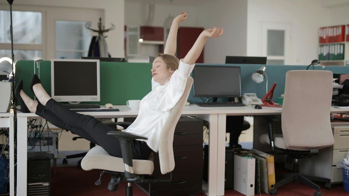 Side view of cheerful female employee in formal outfit sitting on office chair with crossed legs on desk and stretching while resting during work with closed eyes