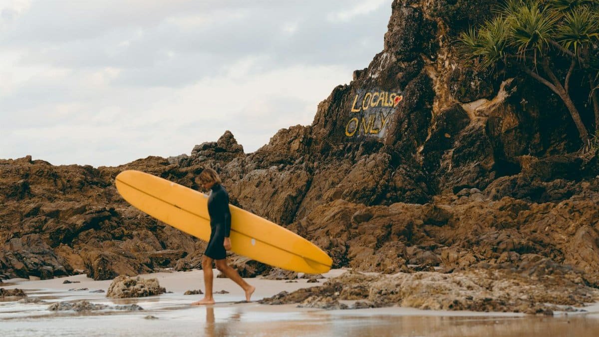 A surfer walks barefoot with a yellow surfboard on the rocky beach of Byron Bay, Australia.
