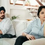 A couple sitting on a couch indoors, looking away from each other, indicating tension.