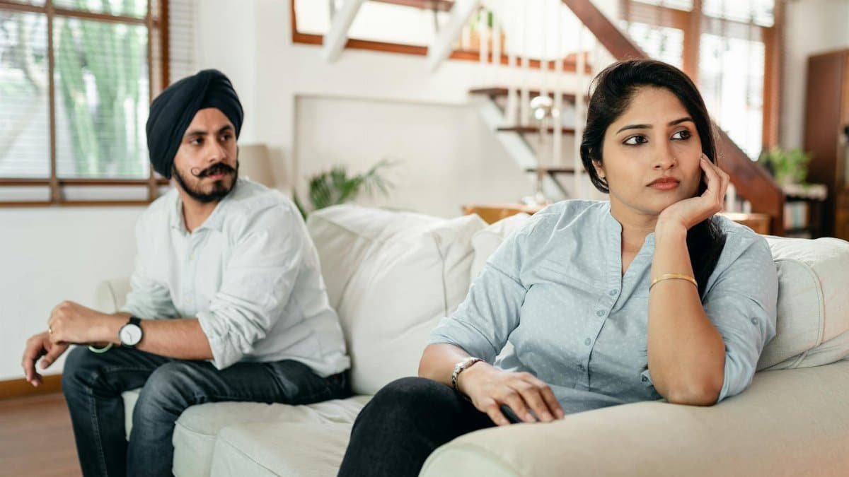 A couple sitting on a couch indoors, looking away from each other, indicating tension.