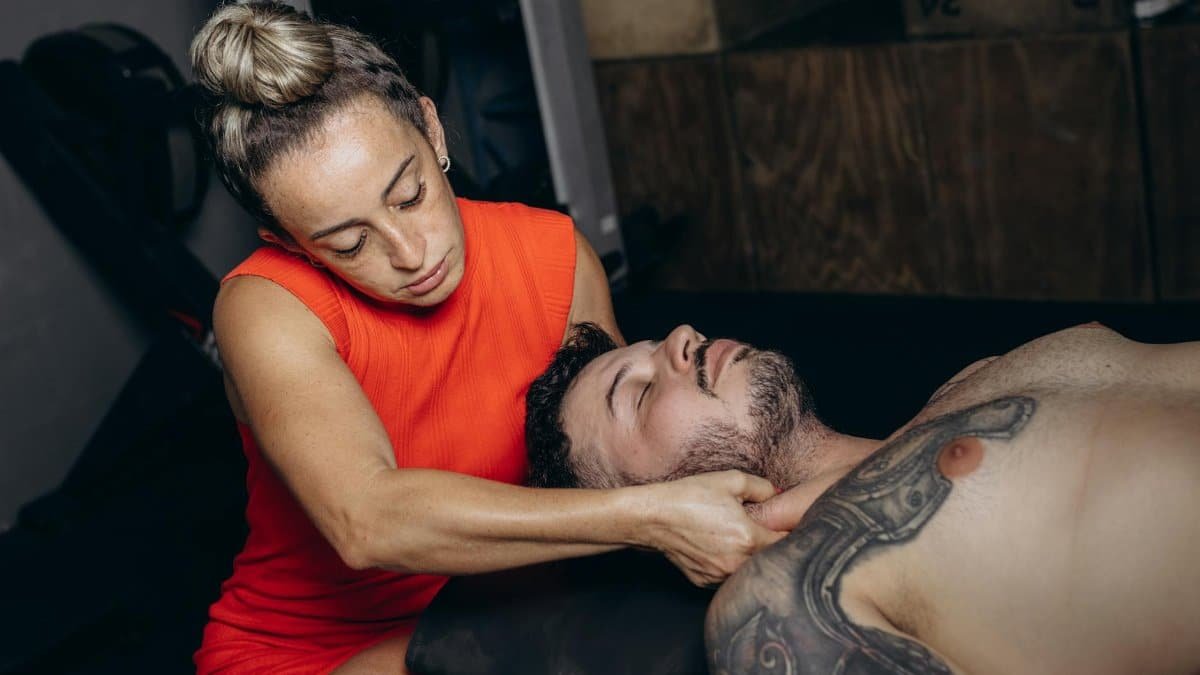 Woman performing therapeutic massage on man's neck in a gym setting.