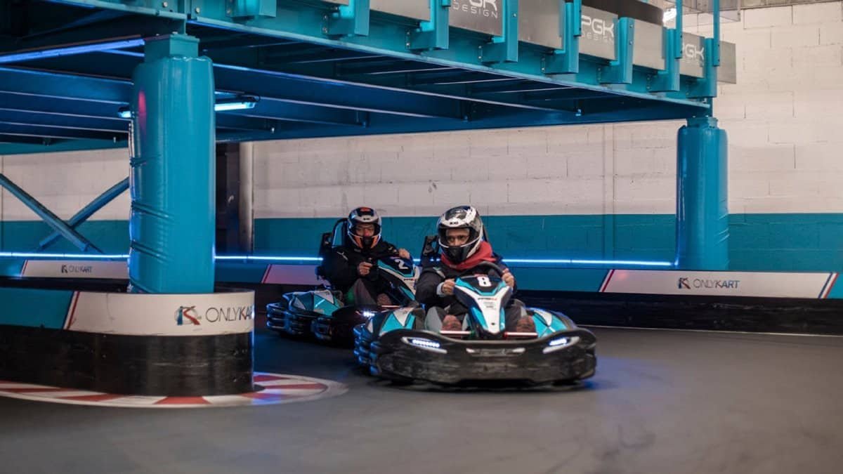 Intense indoor go-kart racing competition with racers maneuvering a sharp turn on the track.